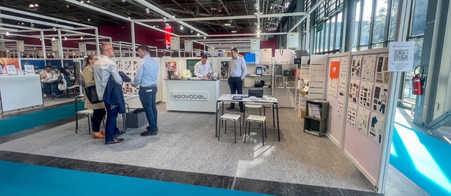 Trade show booth with visitors reviewing printed branding materials at a display table, with product boards and marketing samples showcased in the background.